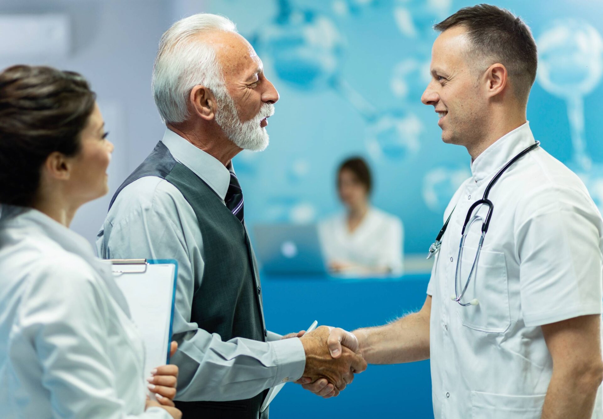 A doctor shakes hands with senior person while greeting in hallway of the clinic accompanied by nurse holding the writing pad 